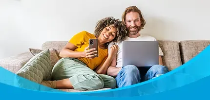 A smiling couple relaxes on a couch sharing a laptop and smartphone together.